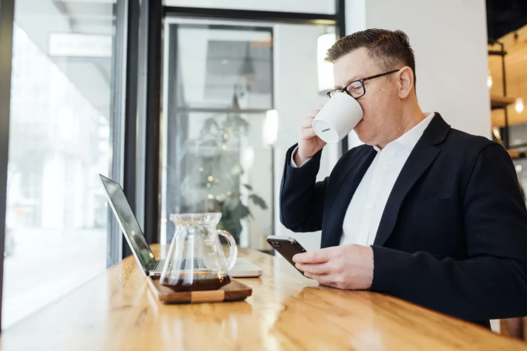 فردی که در محل کار خود پشت لپ تاپ نشسته است و درحال نوشیدن قهوه میباشد و بسیار خندان و خوشحال میباشد و با تلفن همراه خود نیز درحال کار کردن میباشد