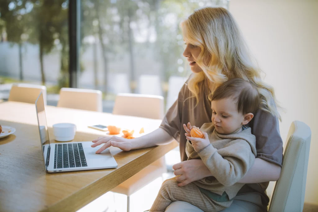 مادر و فرزندی که روی صندلی پای میز نشسته است و مادر درحال کار با لپ تاپ میباشد و فرزند نیز درحال خودن میوه میباشد
