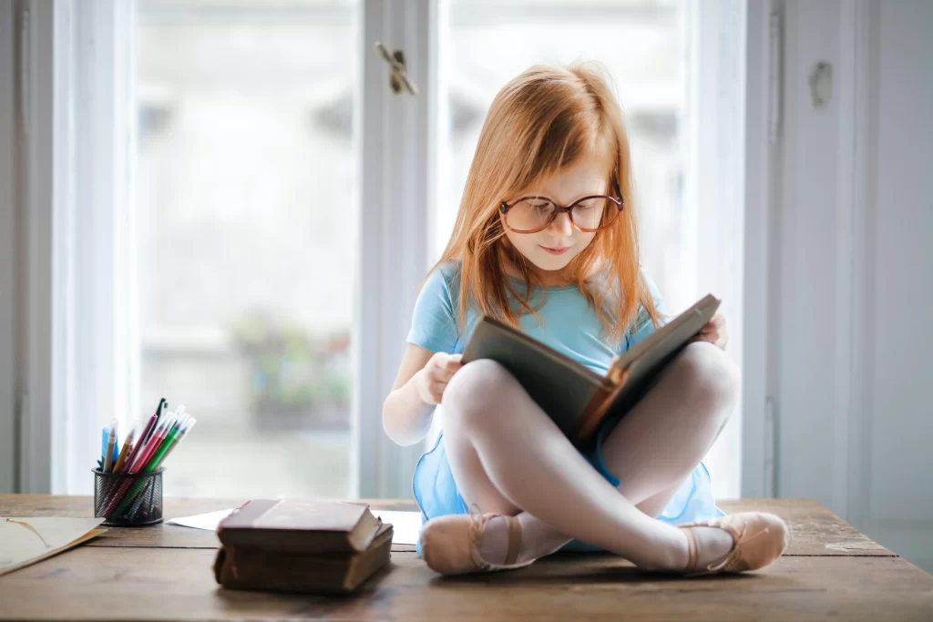 دختر بچه ای که بر روی میز در کنار پنجره نشسته است و درحال خواندن کتاب میباشد و خودکار ها و کتاب نیز در کنارش قرار دارد
