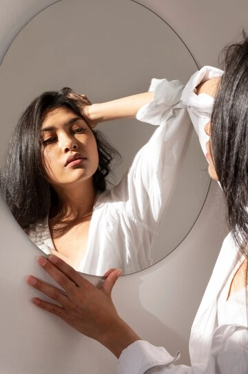 زن جوان با لباس سفید در حال نگاه کردن به خود در آینه بیضی بزرگ روی دیوار